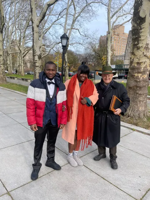 Three people standing on a tree-lined sidewalk, wearing winter coats and scarves, posing outdoors.