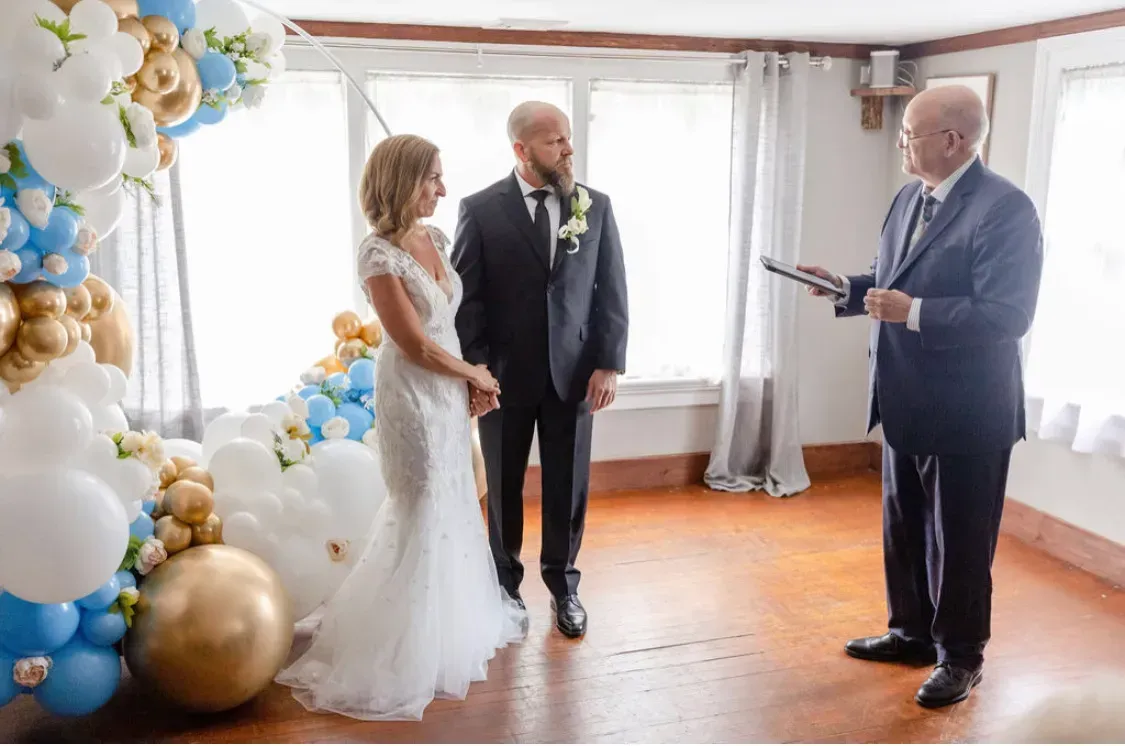 Wedding ceremony indoors with bride, groom, and officiant beside blue, gold, and white balloons
