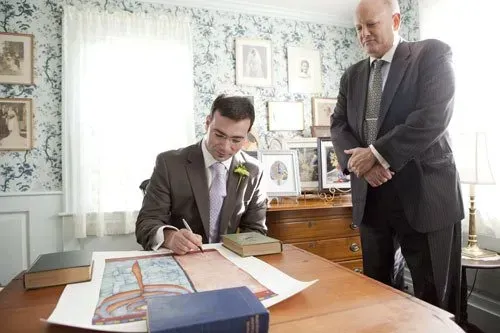 Two men in suits review documents at a desk in a bright office.