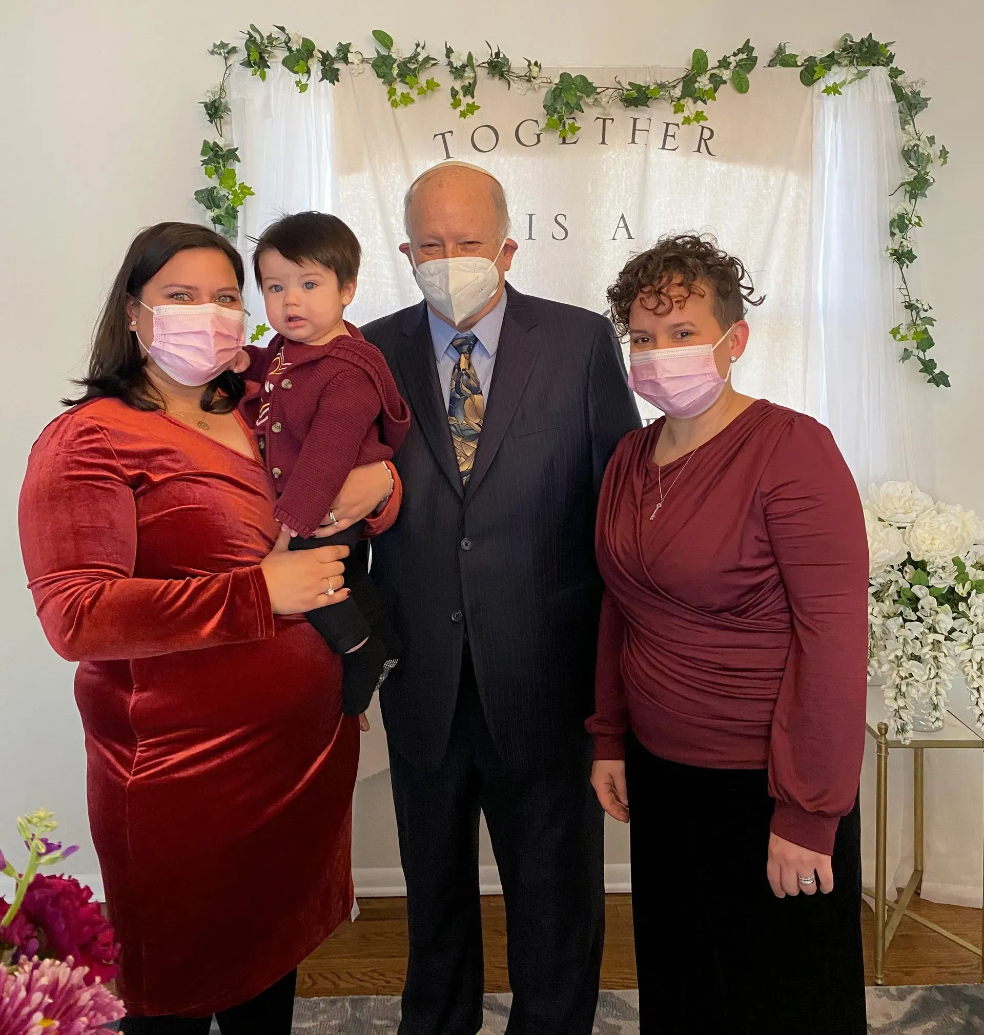 Four people posing indoors at a decorated event, with greenery arch and white backdrop.