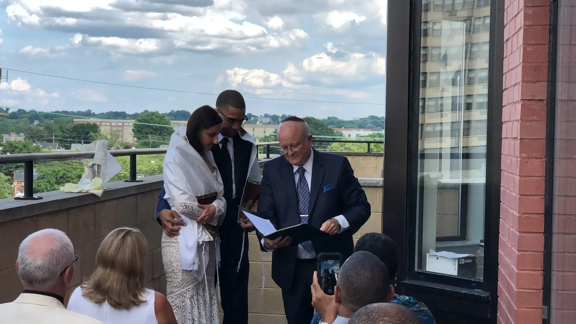 Speaker at a balcony ceremony signs a document while standing with a woman, overlooking a crowd.