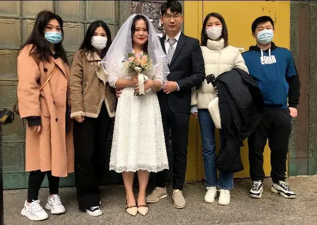 Wedding party posing outdoors, with the bride in a white dress and groom in a black suit centered among four guests.