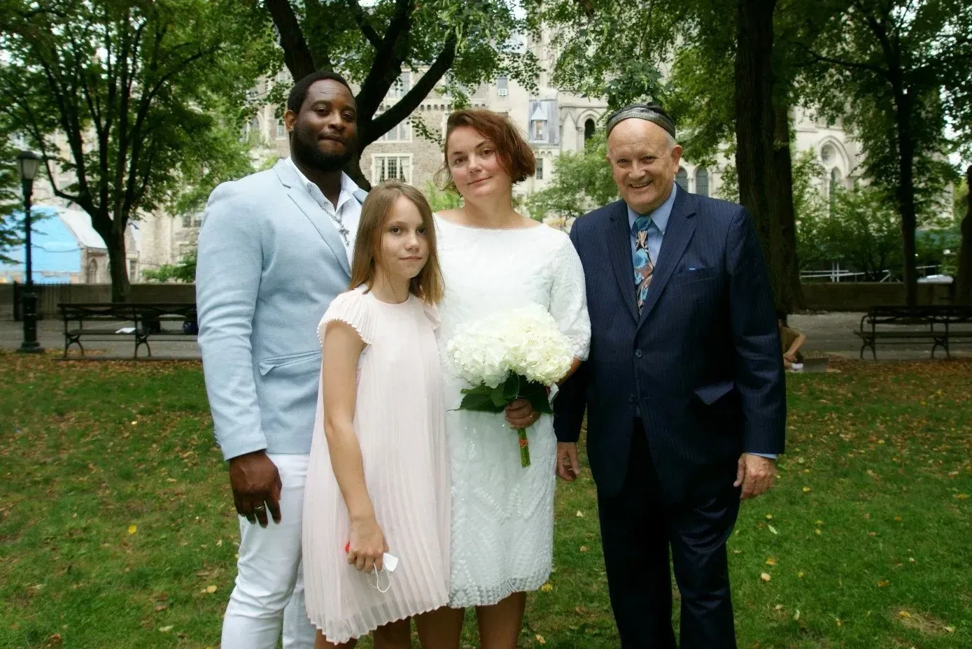Four people posing outdoors in a park, with the center pair holding a white bouquet.