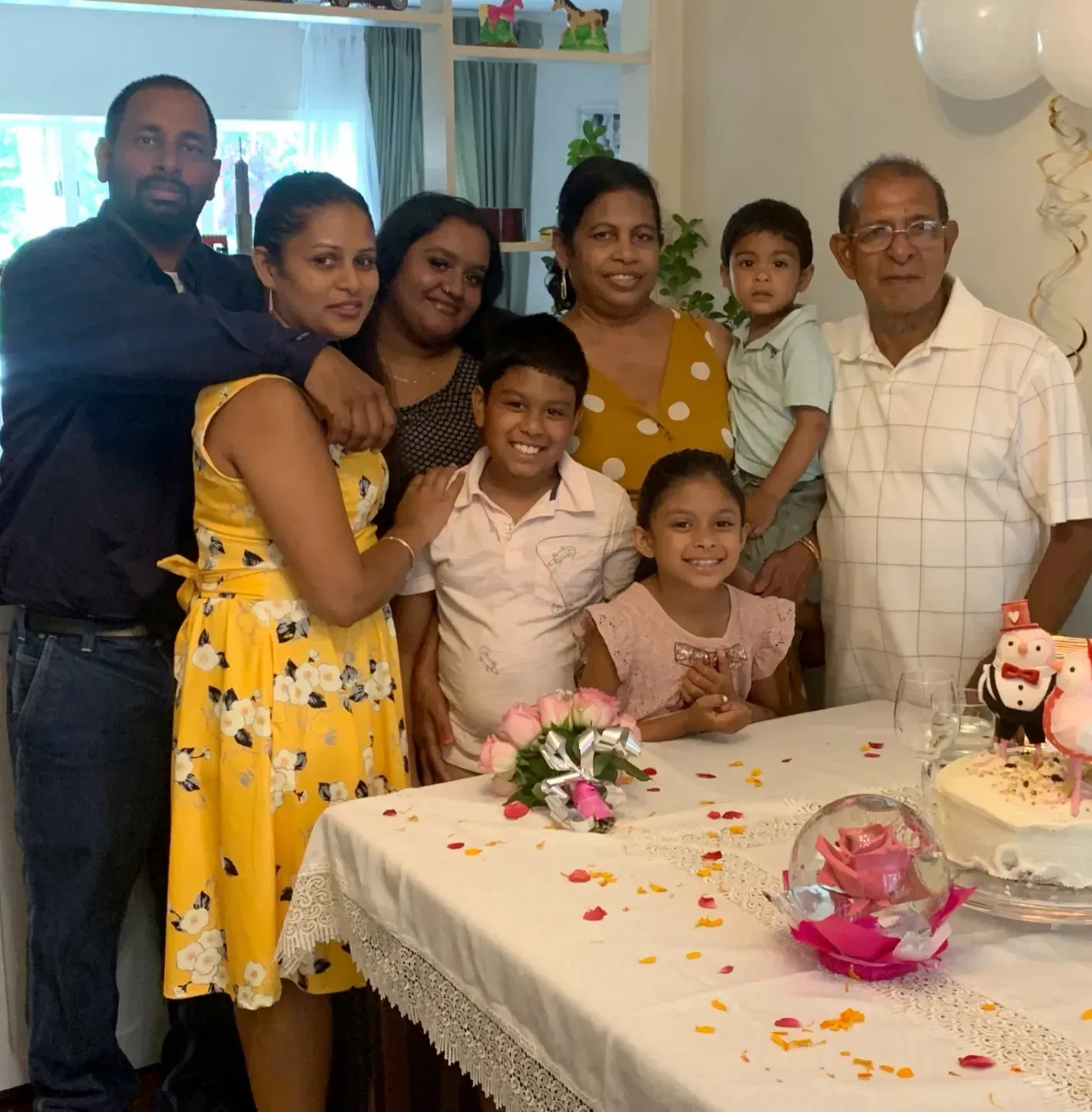 Family gathered around a birthday table with cake, flowers, and balloons, smiling indoors.