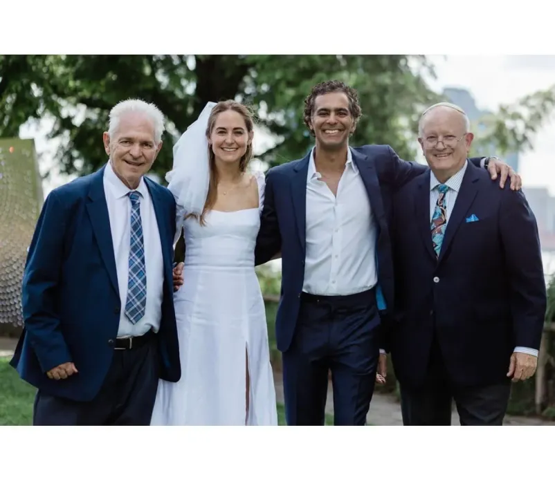 Four people smiling outdoors at a wedding, with two in suits and one woman in a white dress