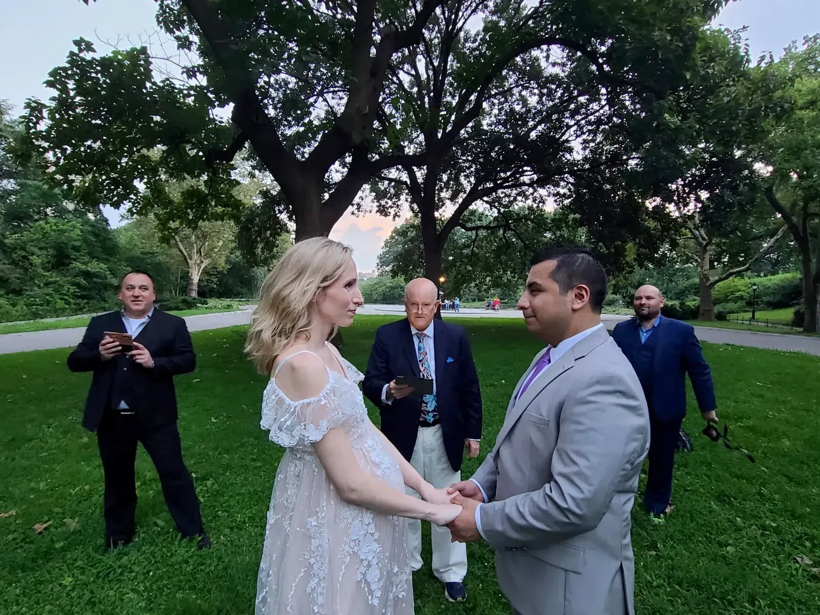 Bride and groom holding hands during an outdoor wedding ceremony in a park with guests behind them