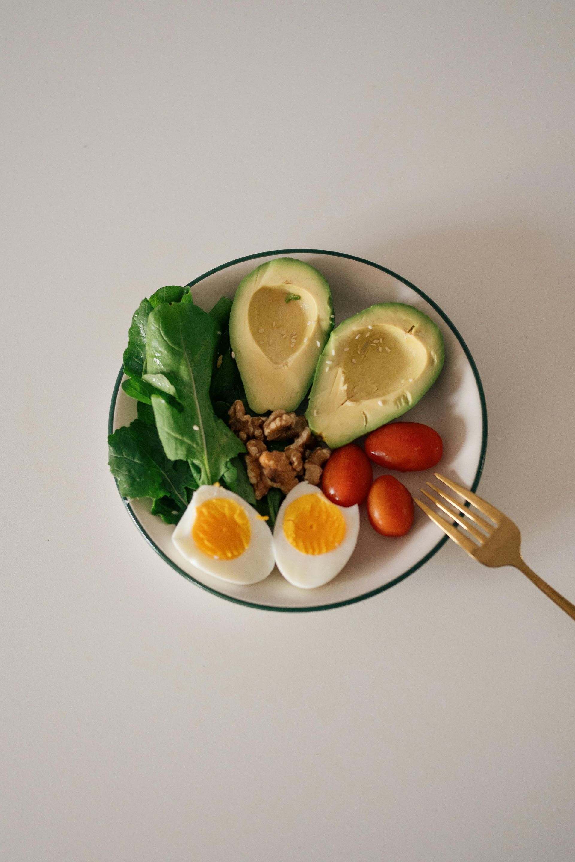 Salad bowl with avocado, eggs, greens, cherry tomatoes, walnuts, and a fork on a light table