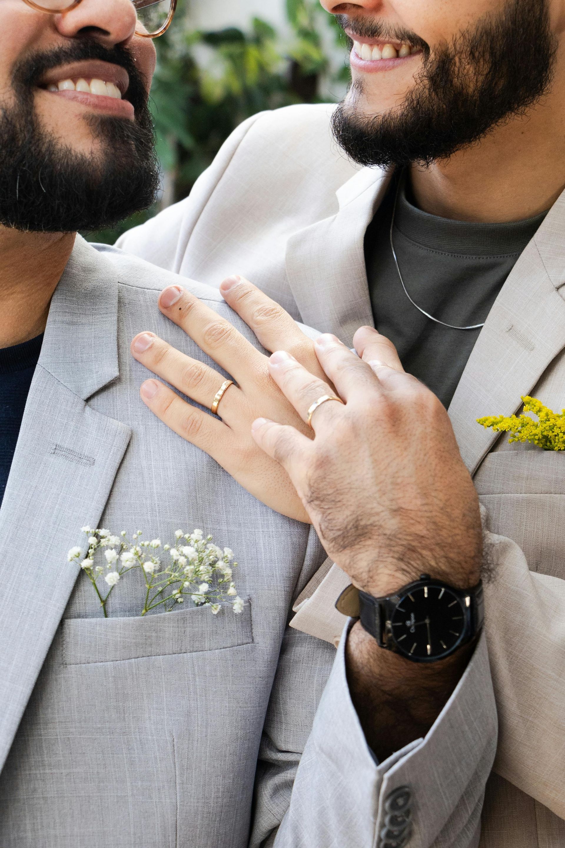 Two men in light suits smiling closely, one showing a hand with wedding rings.