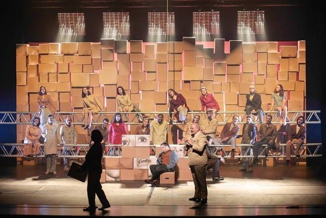 Actors perform on a stage set of stacked cardboard boxes with warm lighting.