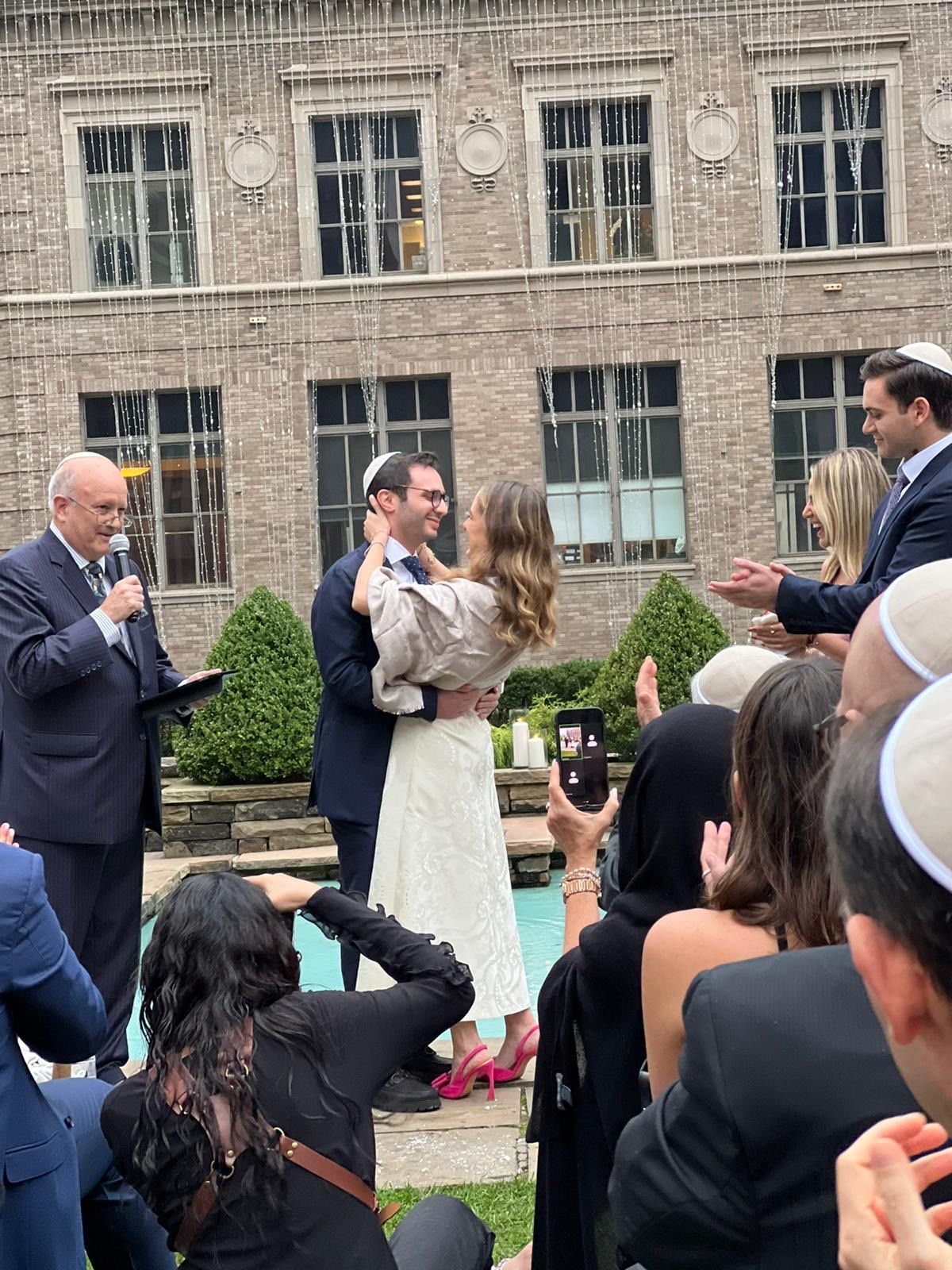 Outdoor wedding ceremony with couple at center, officiant reading, and guests gathered in front of a stone building