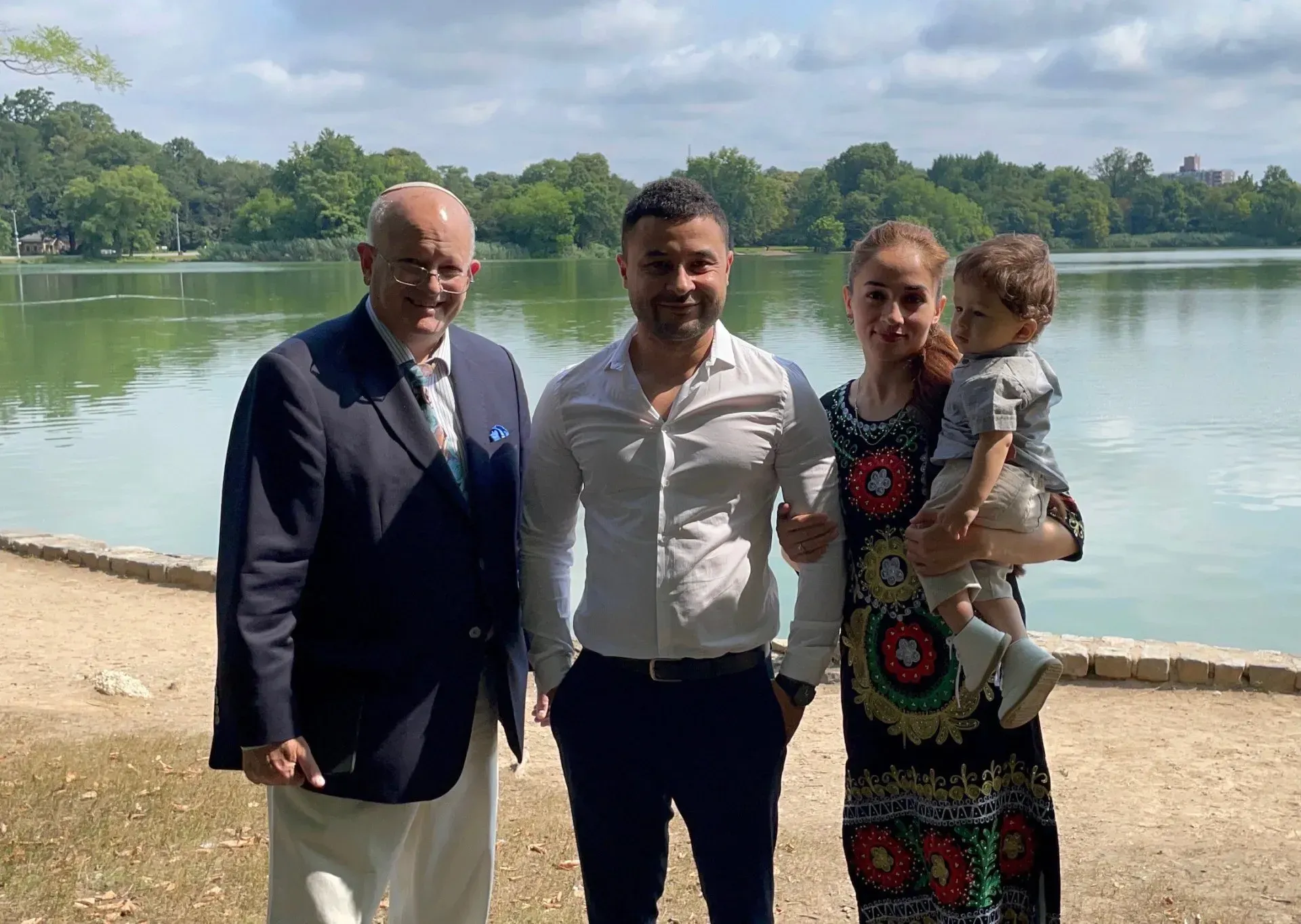 Four people posing by a lake on a sunny day, with trees and water in the background.