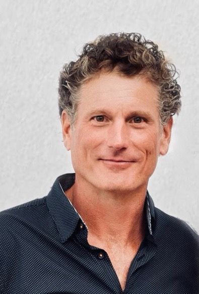 Portrait of a smiling person with curly hair wearing a dark blue shirt against a light gray background