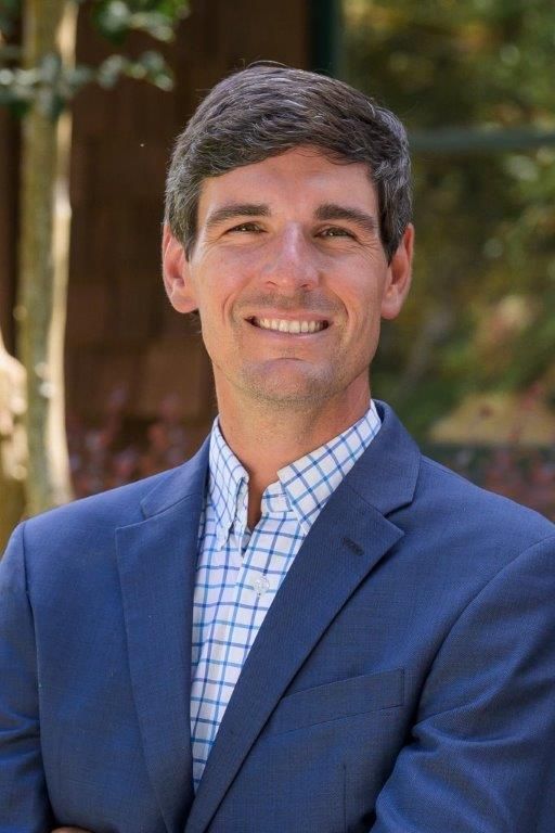Smiling man in a blue blazer and checkered shirt outdoors, with blurred trees behind him.