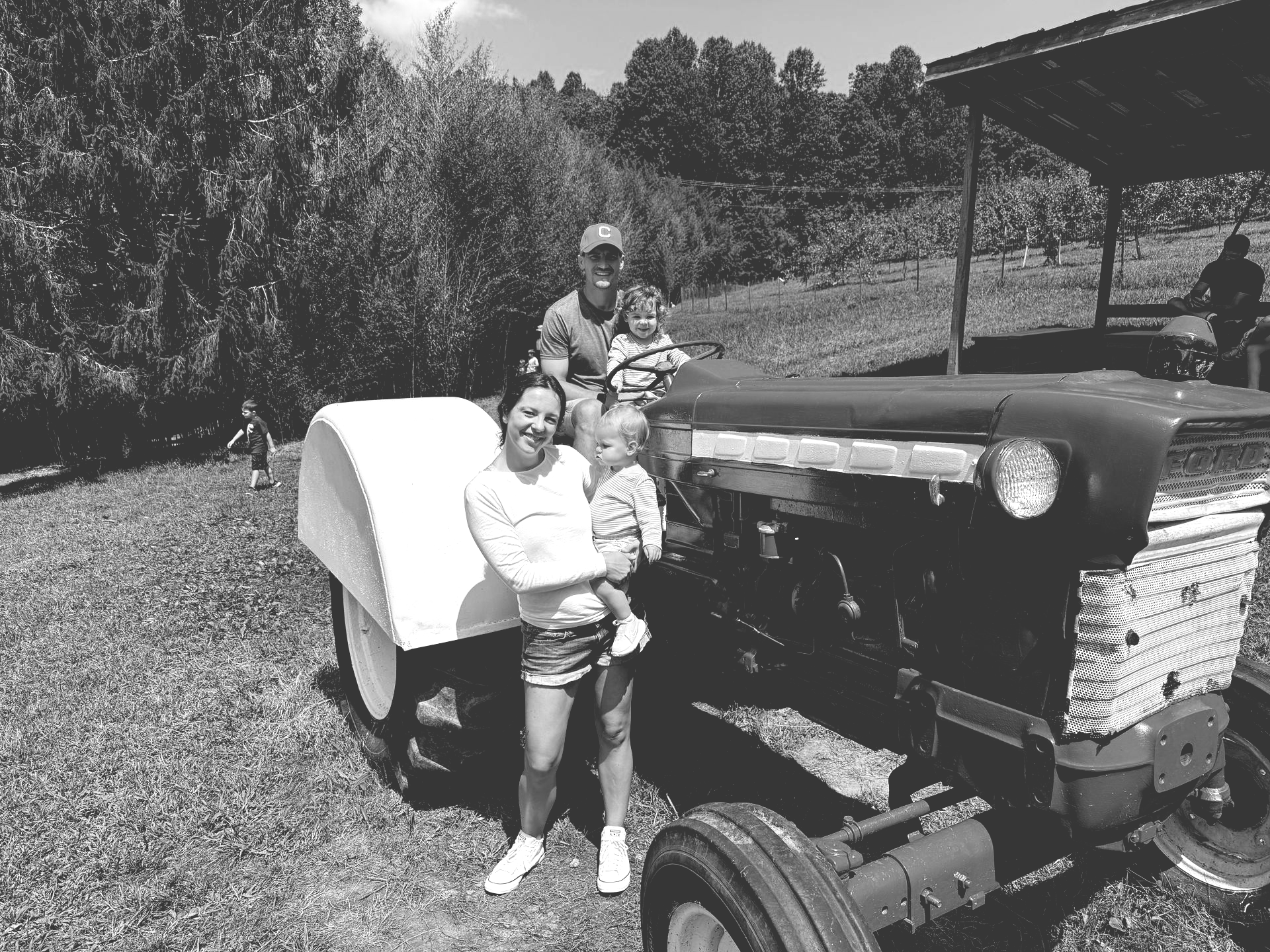 Two people pose beside a vintage tractor with a covered trailer on a grassy farm field.