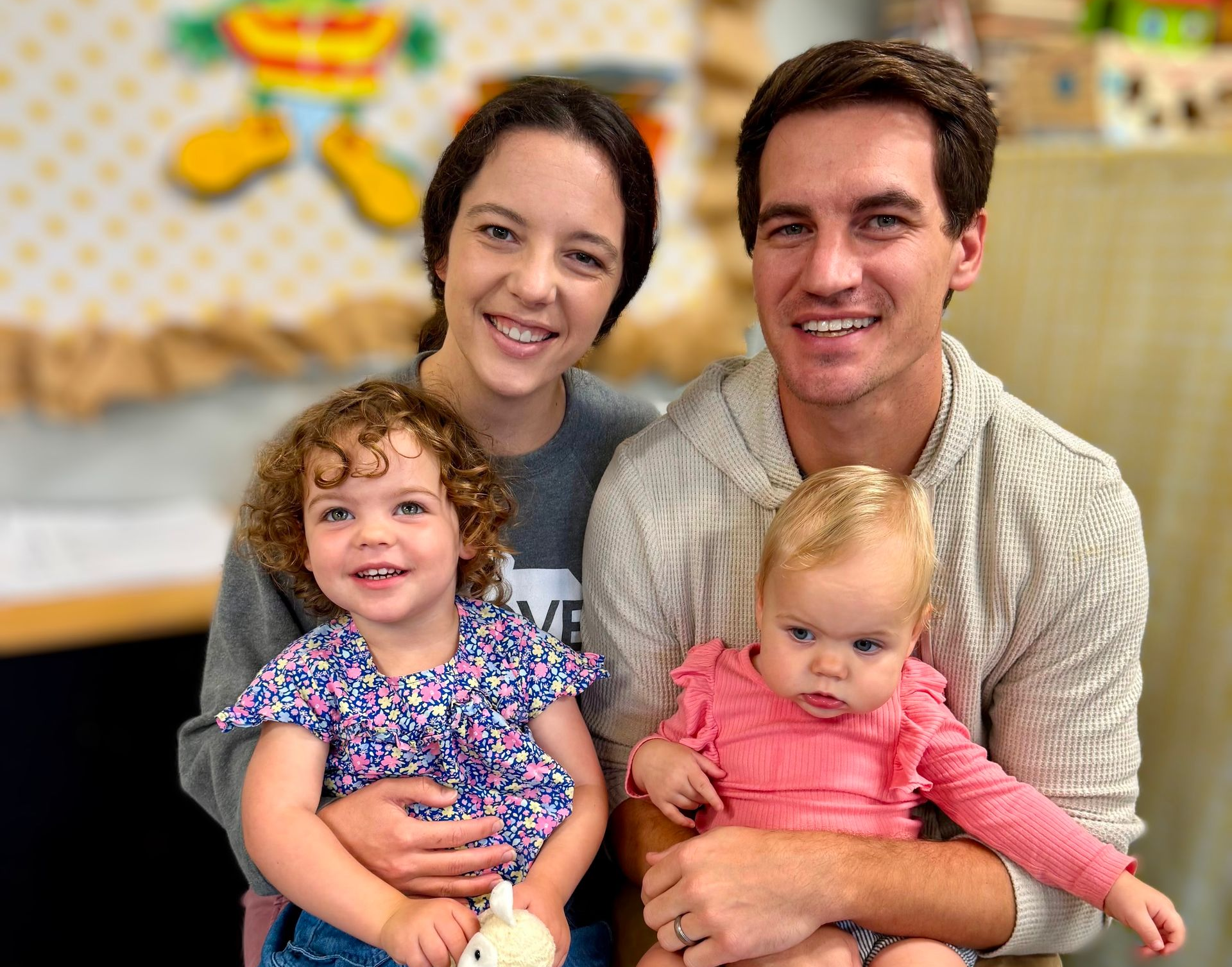 Two adults holding two smiling toddlers in a bright indoor setting