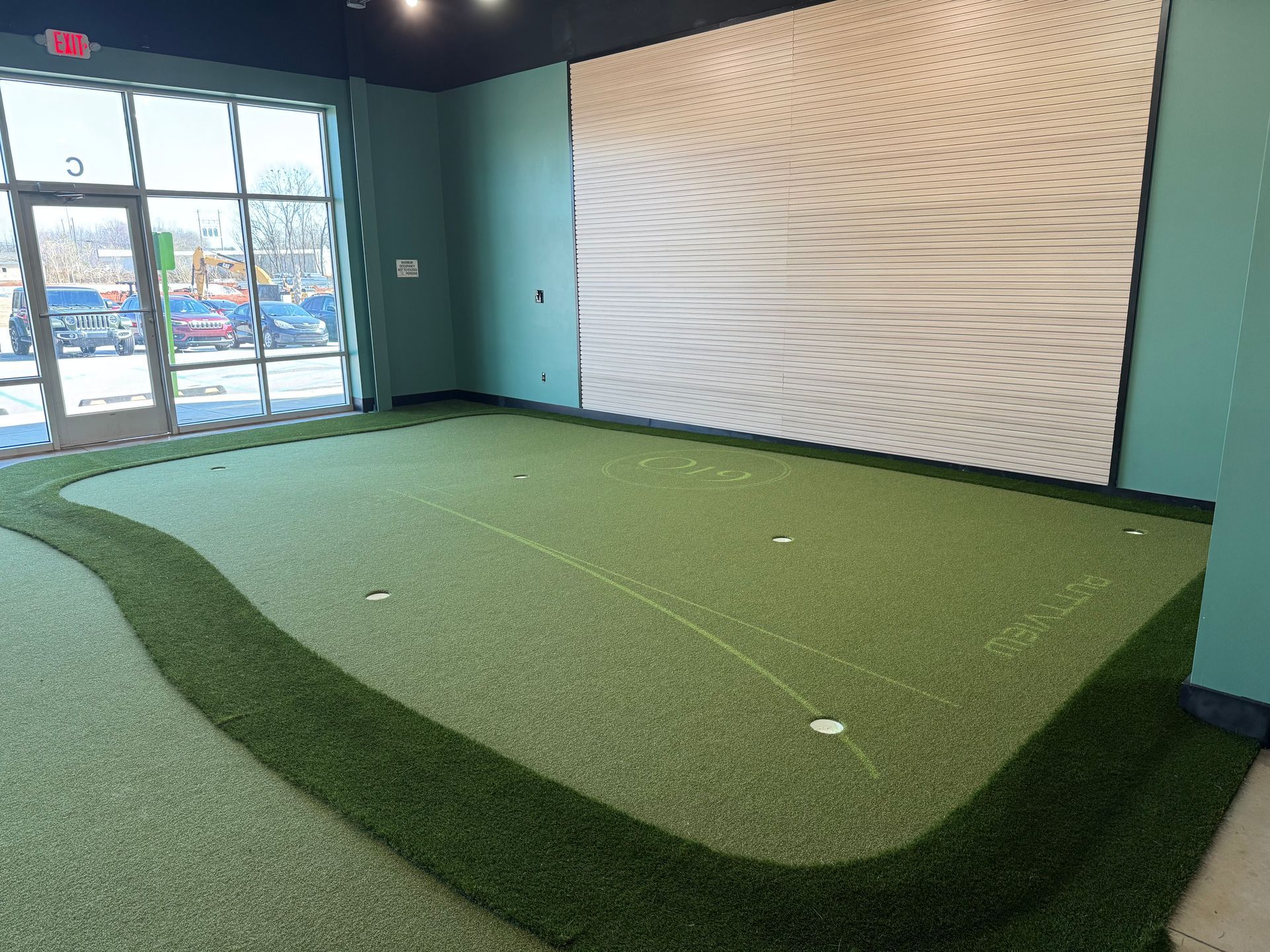 Indoor putting green with artificial turf, near large windows and a white accent wall