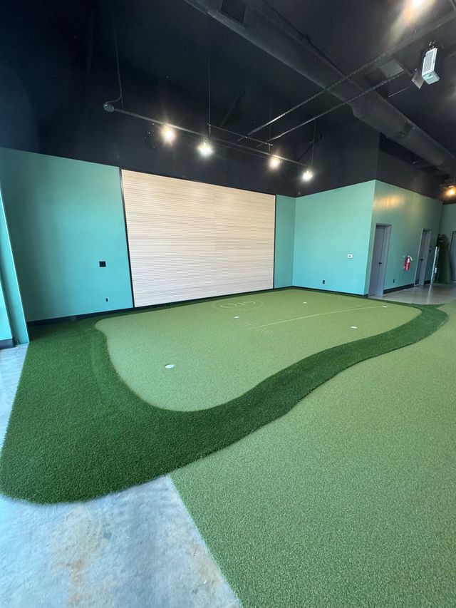 Indoor mini golf putting area with green turf, turquoise walls, and a white target wall under ceiling spotlights