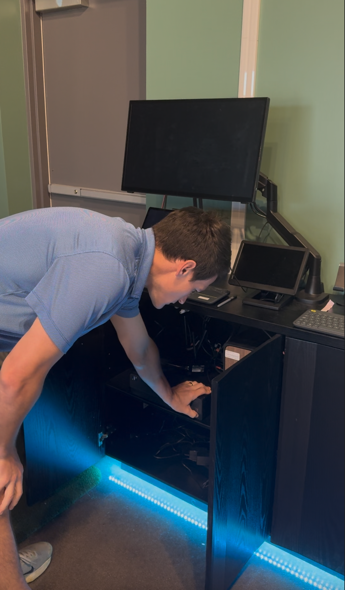 Person bending over to inspect black computer equipment with a glowing blue LED strip in a room.
