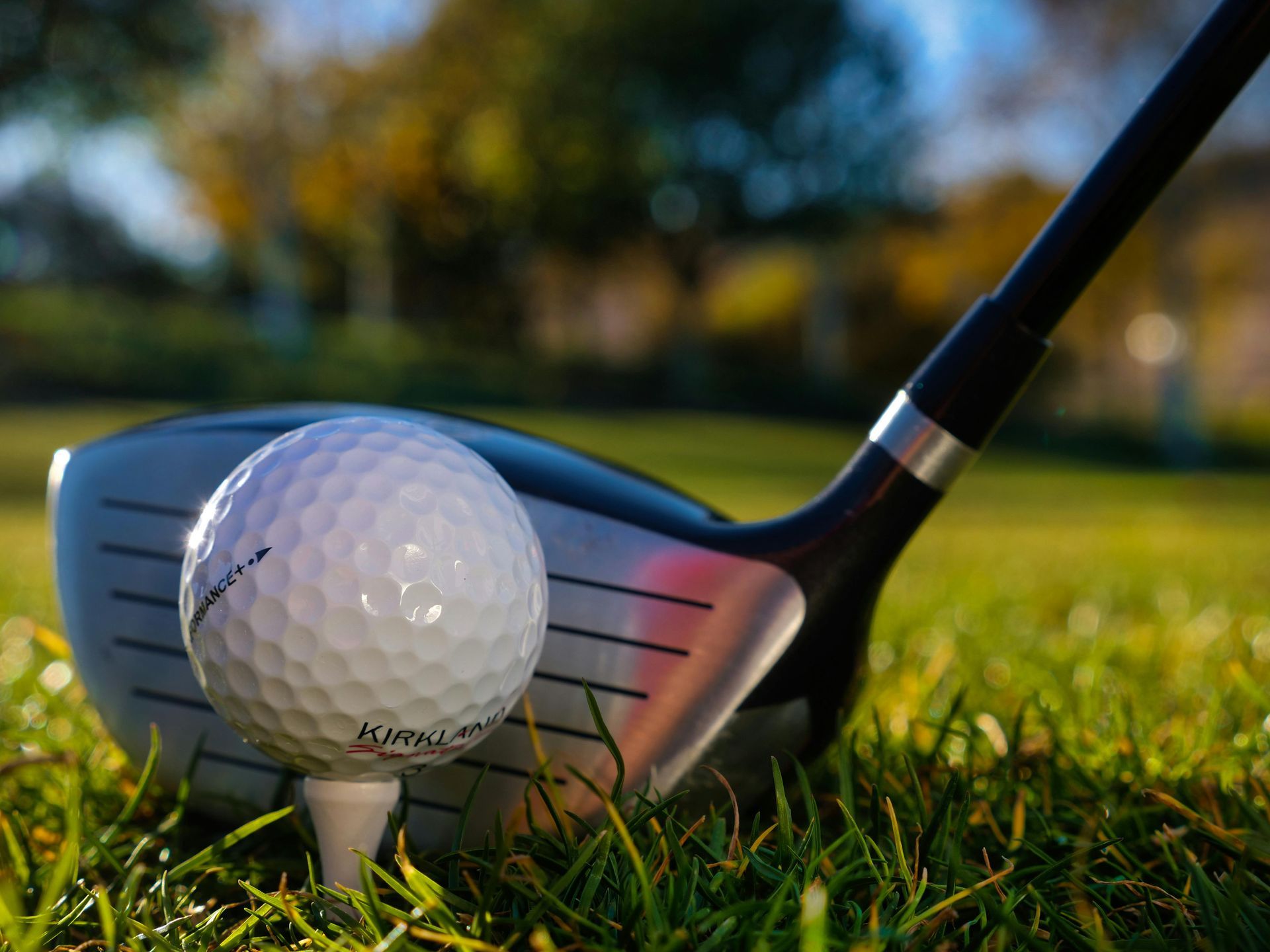 Golf ball on a tee in front of a golf club on a grassy course at sunset
