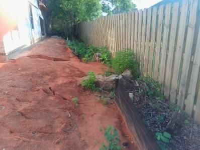 Red dirt path beside a wooden fence and building with sparse green plants.