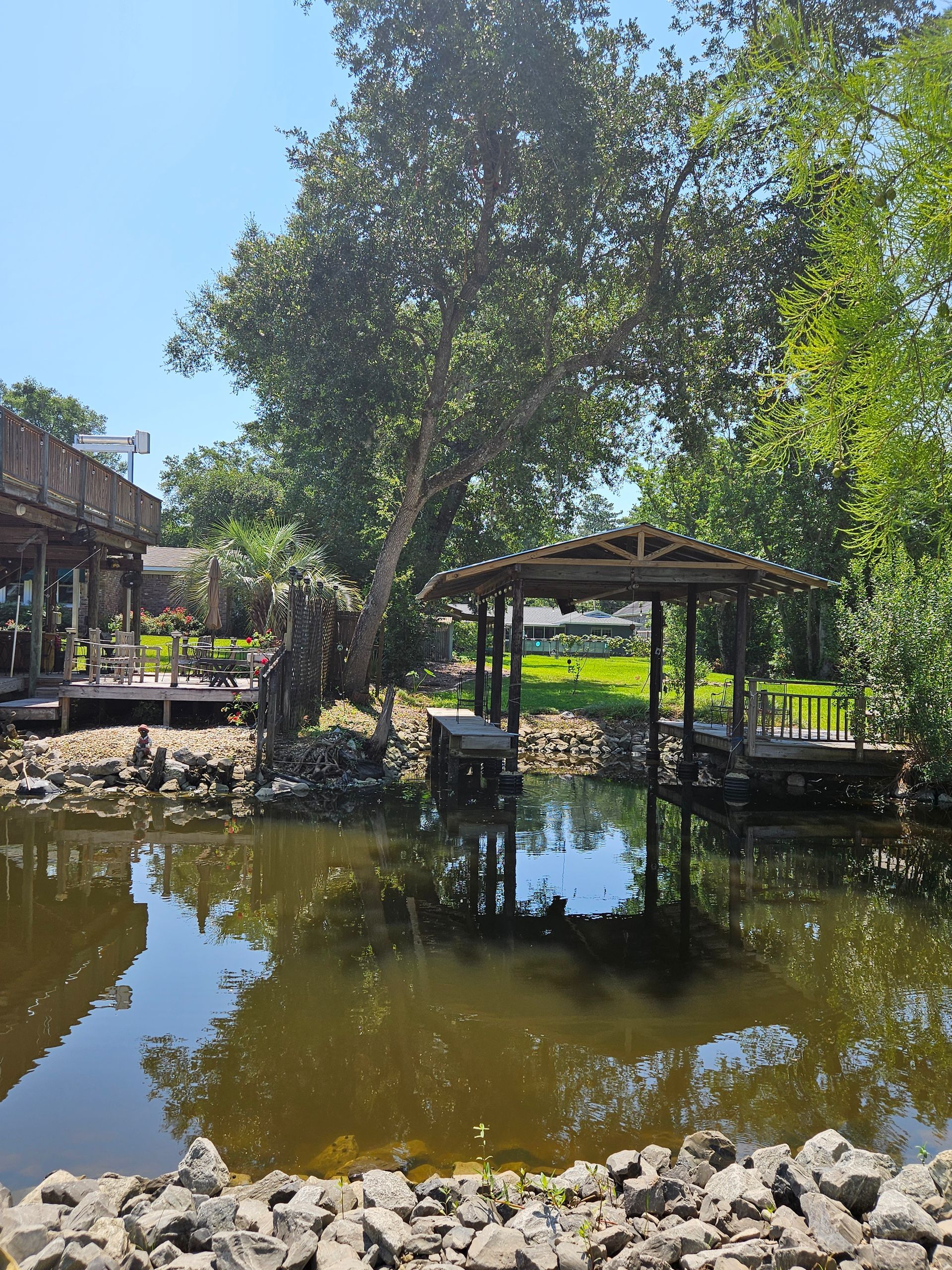 A gazebo and trees reflect in a dark pond, surrounded by rocks and a wooden deck. Sunny day.