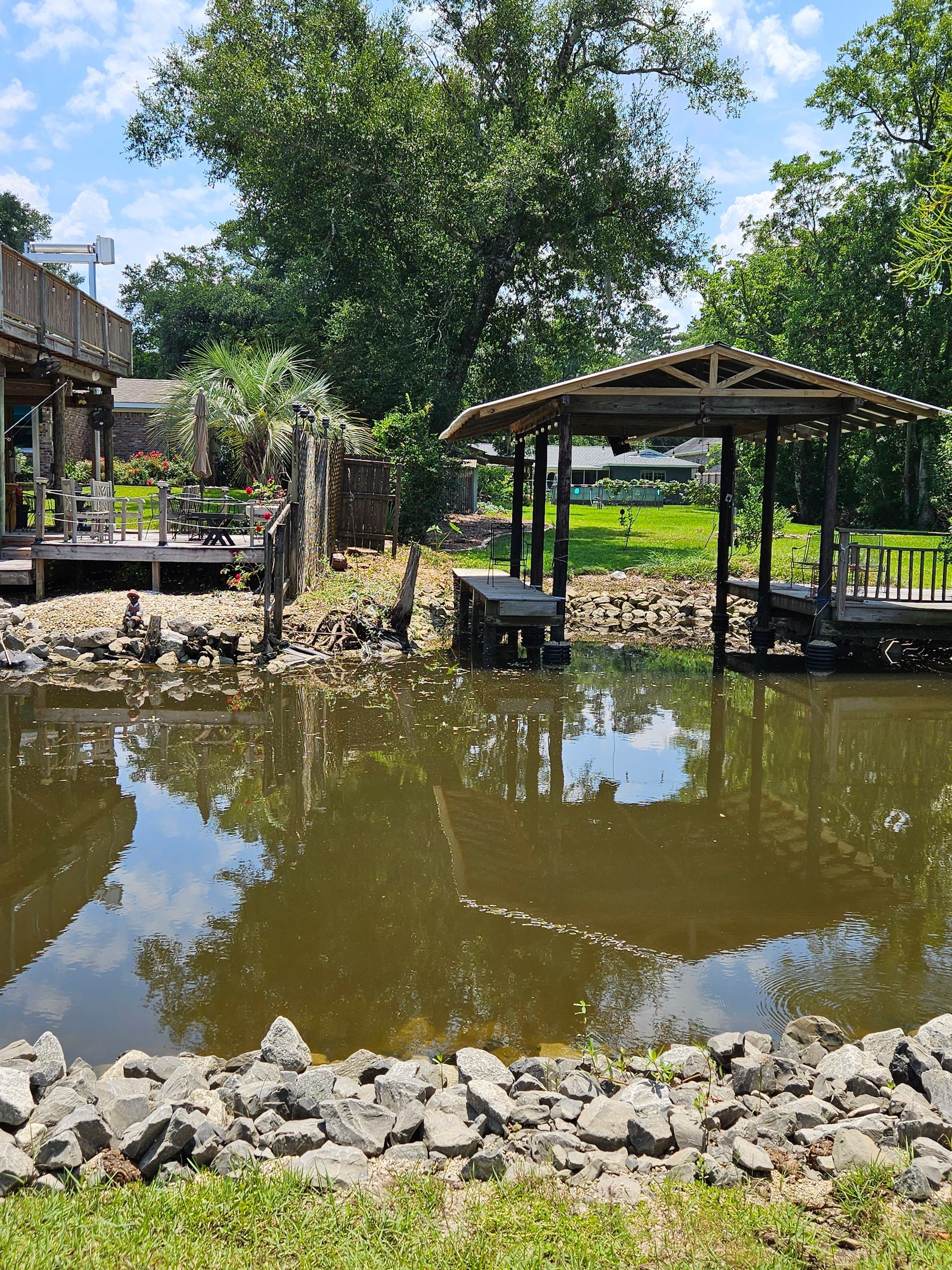 Pond with a wooden gazebo, a deck, and surrounding greenery under a cloudy sky.