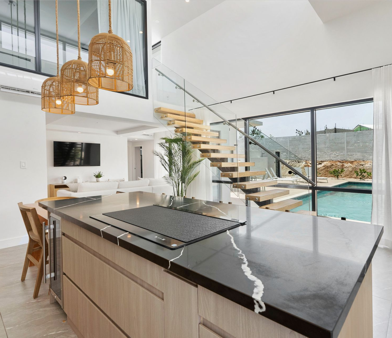 Modern open-plan kitchen with island, hanging pendant lights, and a floating staircase overlooking a pool and hills