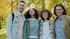 Four friends standing together outdoors in a sunny park, smiling at the camera.