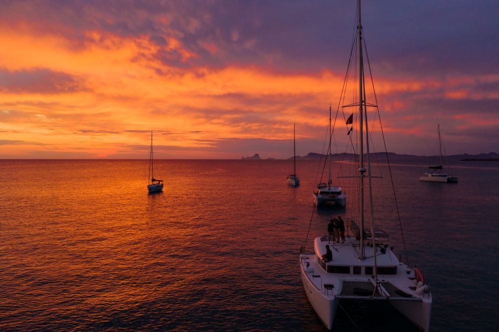 Sailboats at sunset on calm orange water, with a catamaran in the foreground