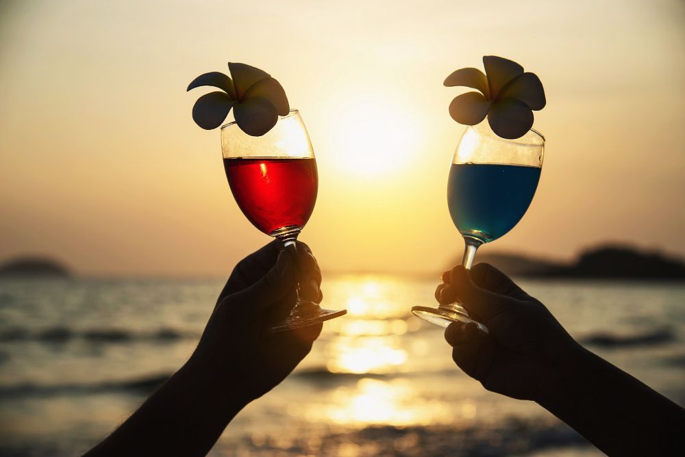 Two hands toasting wine glasses at sunset over the sea, one red drink and one blue