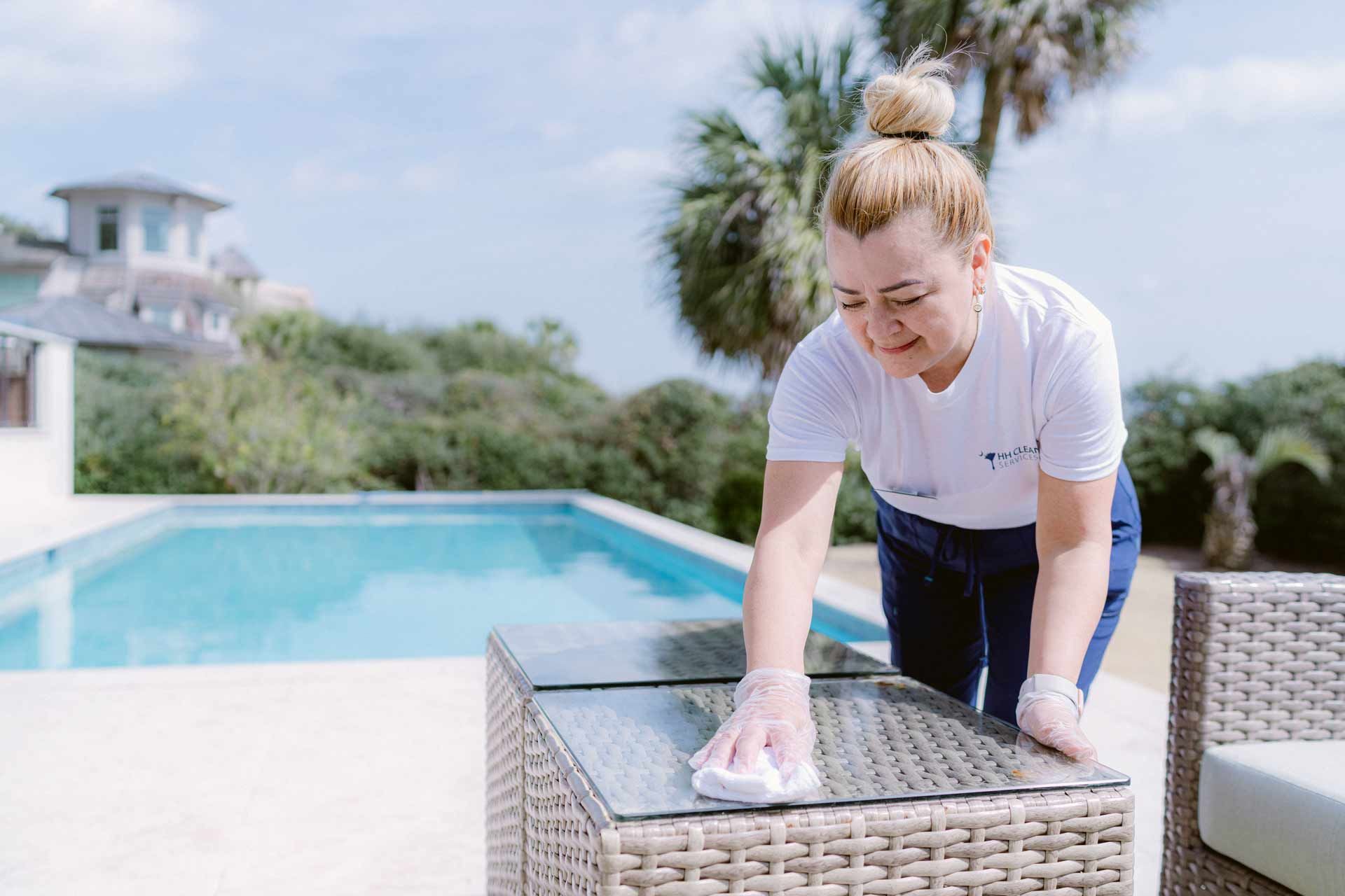 Person wiping a wicker table beside a pool, with palm trees and a bright patio in the background