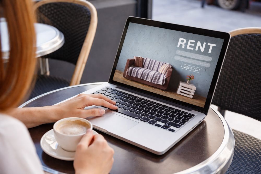 Person using a laptop at a café table, browsing a rent listing with a sofa photo on screen.