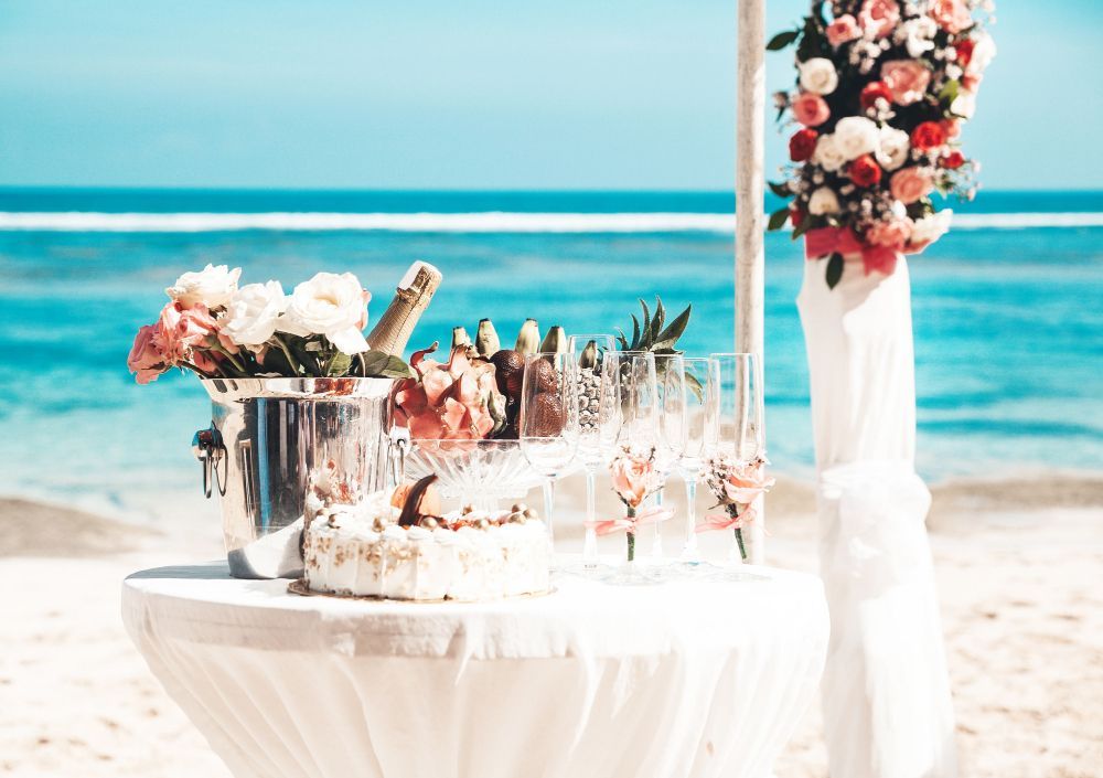 Beachside wedding dessert table with cake, champagne, flowers, and white decor near the ocean