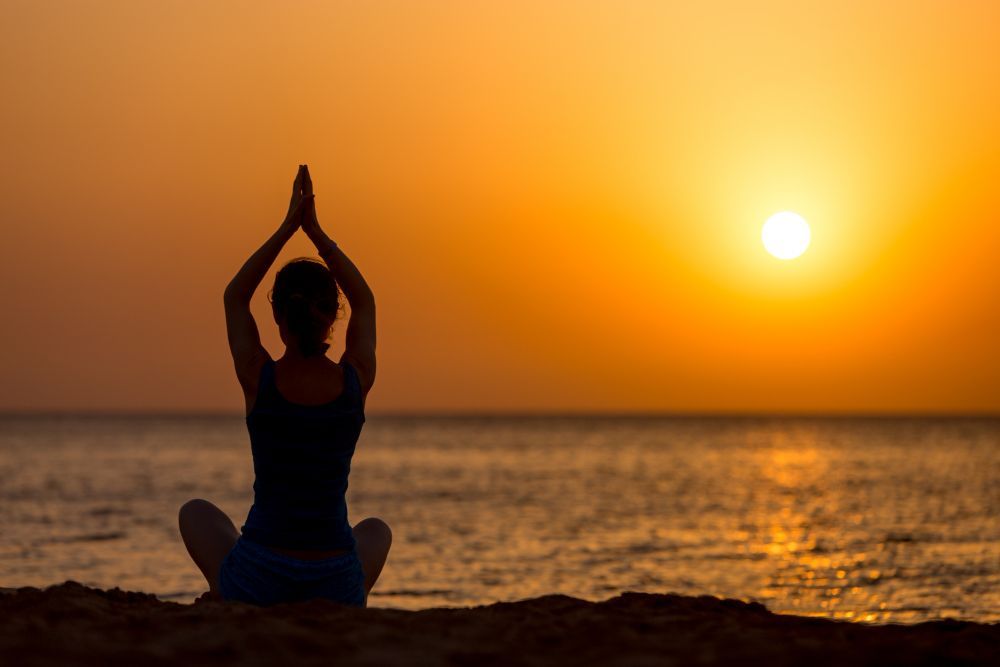 Person doing yoga on a beach at sunset with arms raised toward the glowing sun