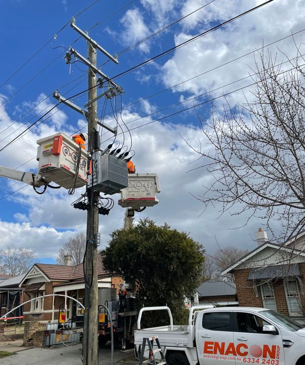 Linemen working on electrical equipment on a utility pole. White bucket trucks, houses, and a blue sky.