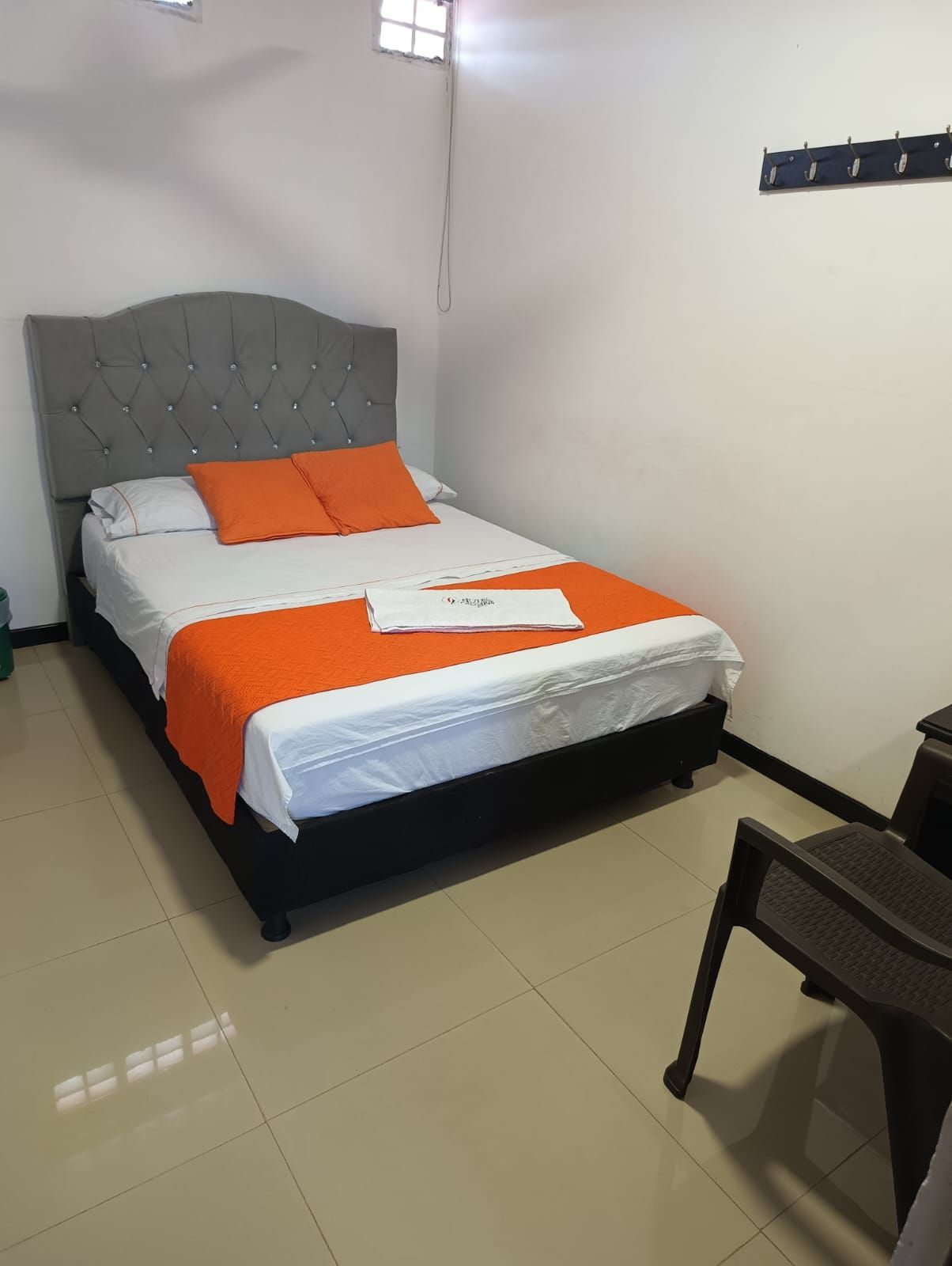 Habitación pequeña con una cama gris, ropa de cama naranja, una estantería en la pared y una silla en primer plano.
