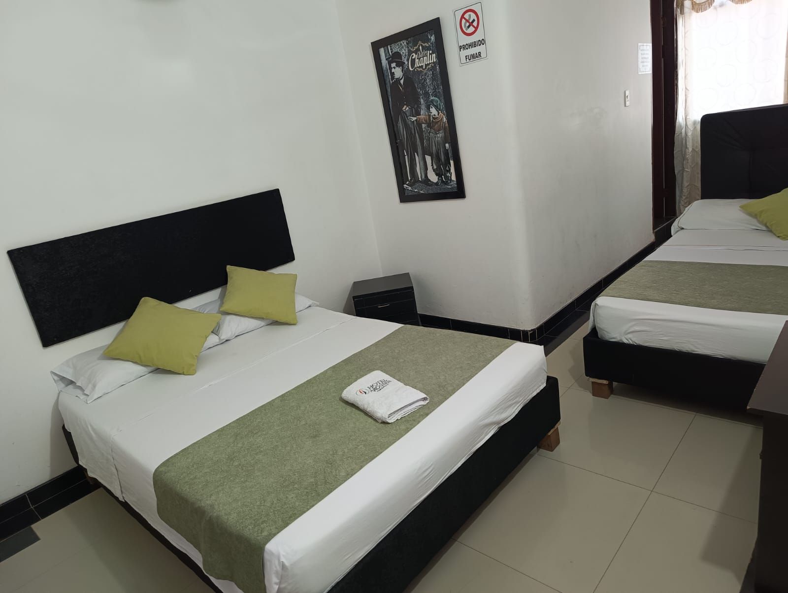Habitación de hotel pequeña con dos camas bien hechas, sábanas blancas, alfombras verdes y suelo de baldosas.
