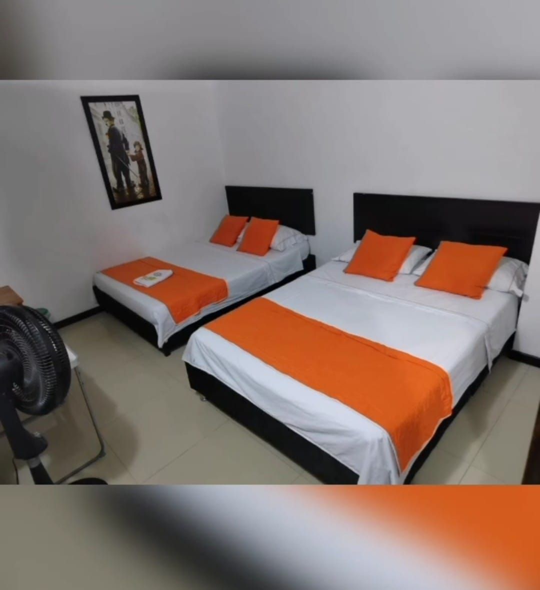 Habitación pequeña con dos camas bien hechas, detalles en color naranja y un cuadro en la pared.