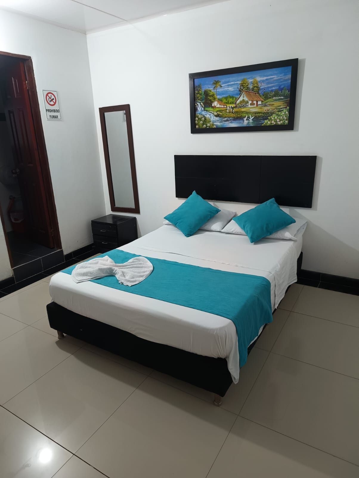 Pequeña habitación de hotel con cama doble, detalles en color turquesa, cuadros en la pared y un televisor