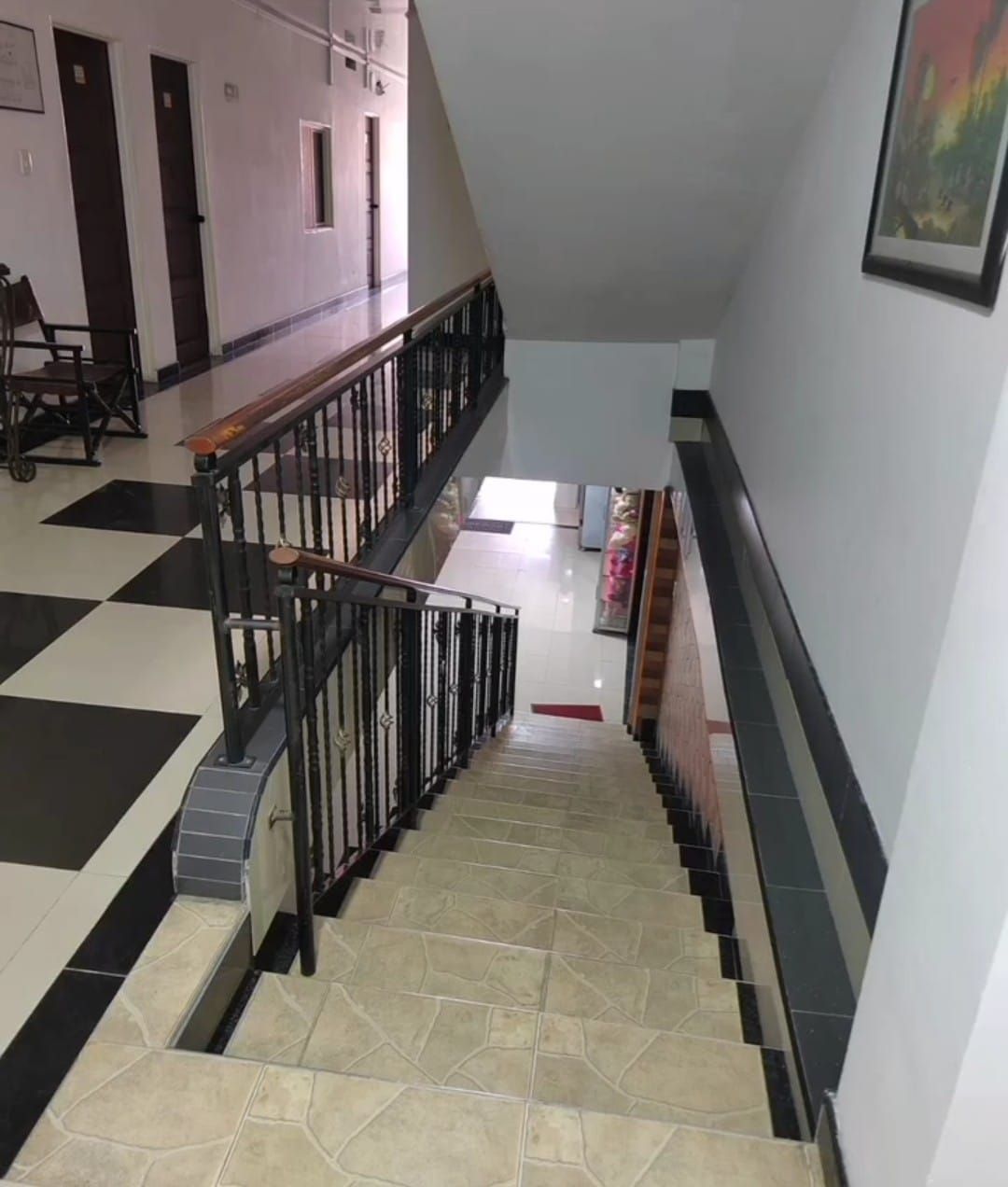 Escalera interior con barandillas negras y baldosas beige, junto a un pasillo con suelo de baldosas a cuadros.