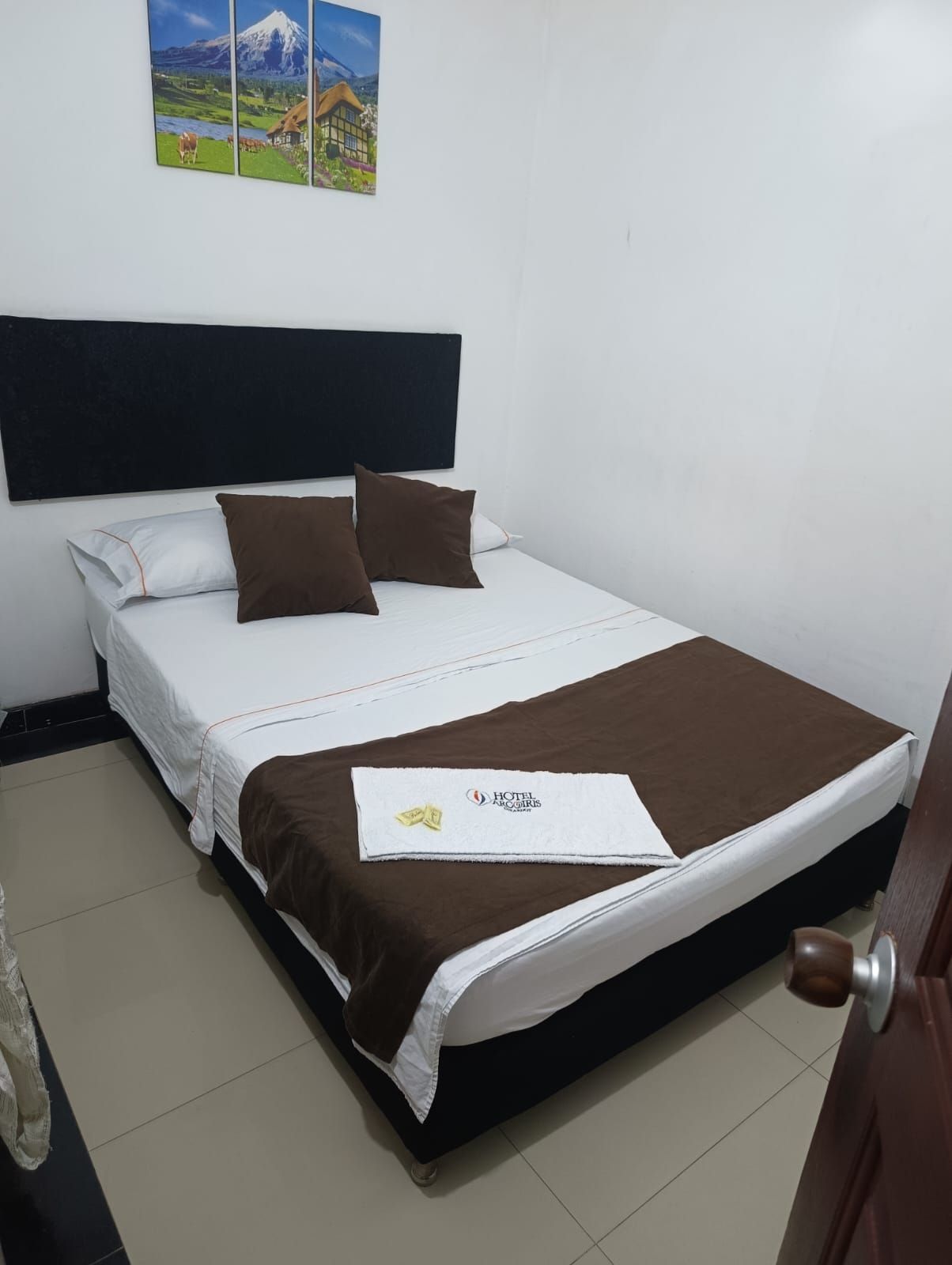 Habitación pequeña con una cama bien hecha, ropa de cama marrón y blanca y un cuadro con un paisaje en la pared.
