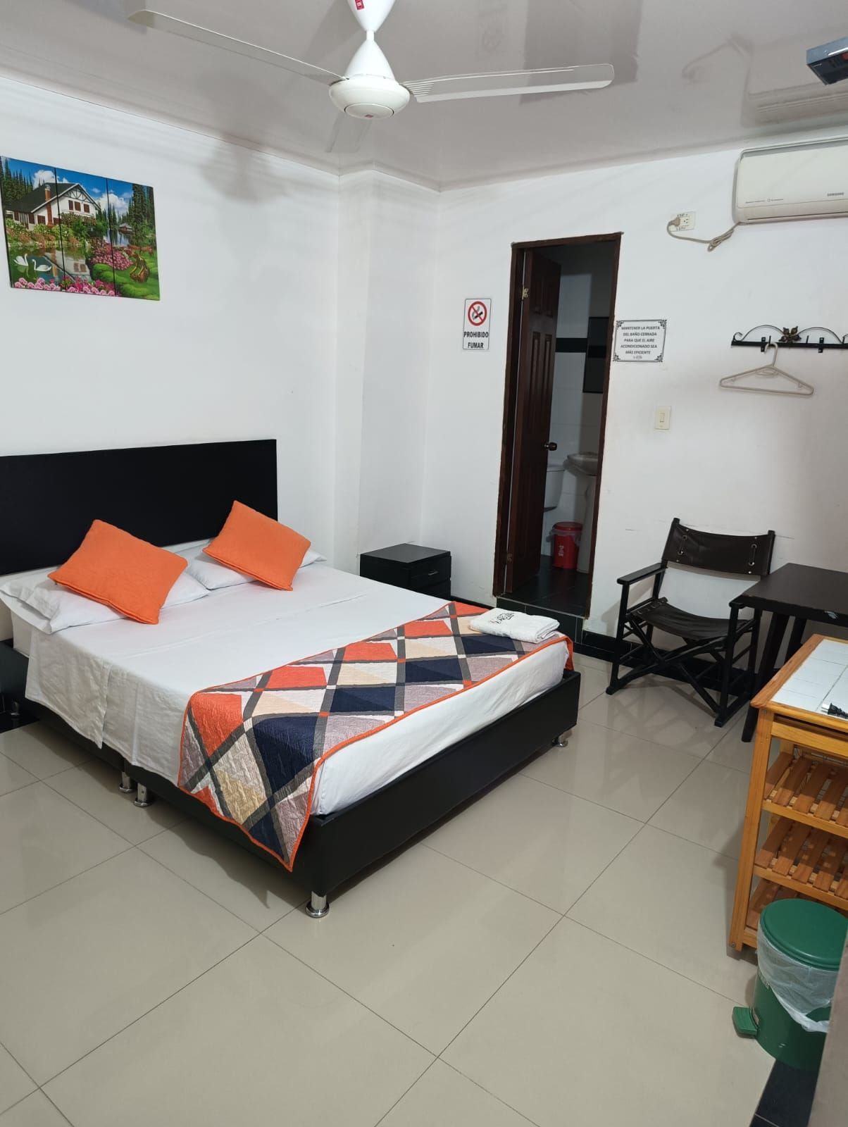 Habitación pequeña con cama, almohadas naranjas, ventilador de techo, silla y unidad de aire acondicionado