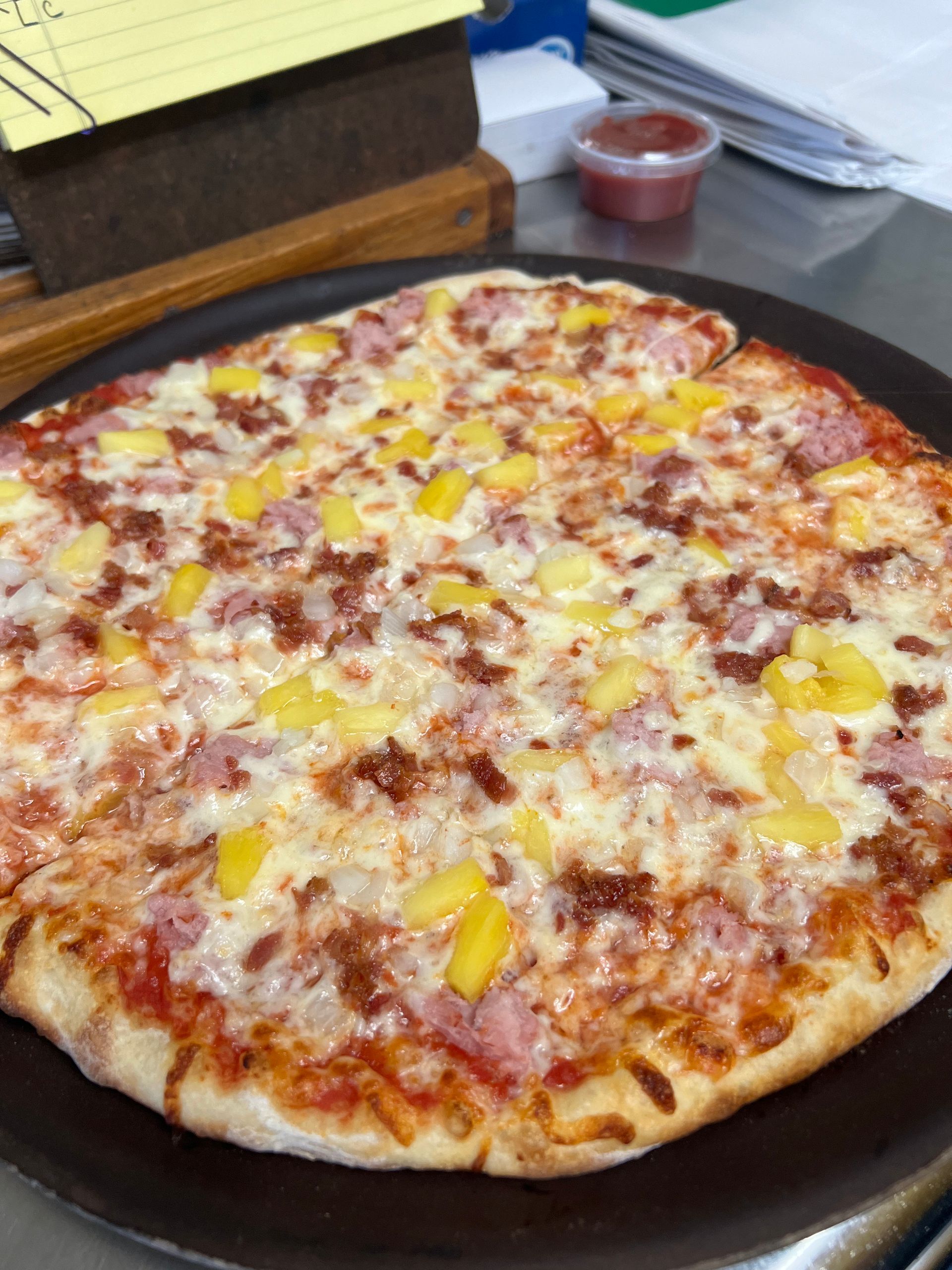 A pizza with ham and pineapple is on a pan on a table.