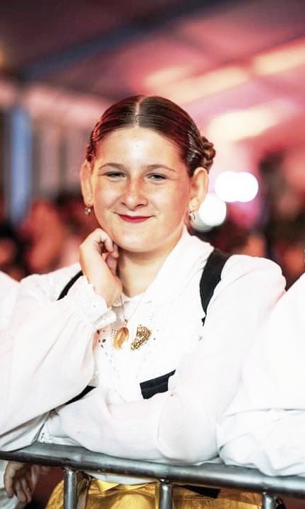 A young girl in a white shirt is leaning against a fence and smiling.