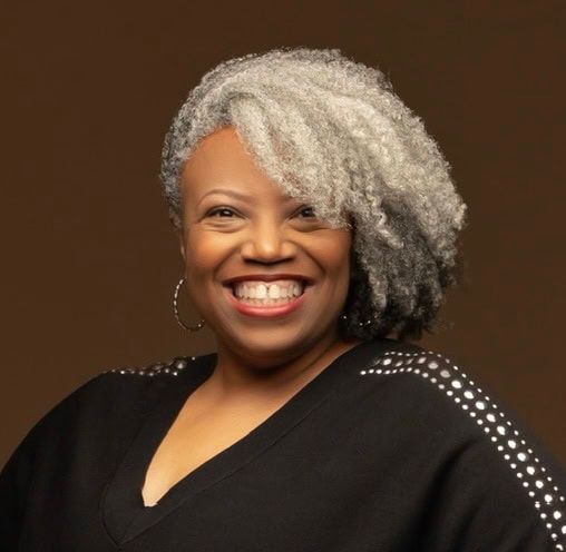 Smiling person with silver-gray hair in a black top against a dark brown background