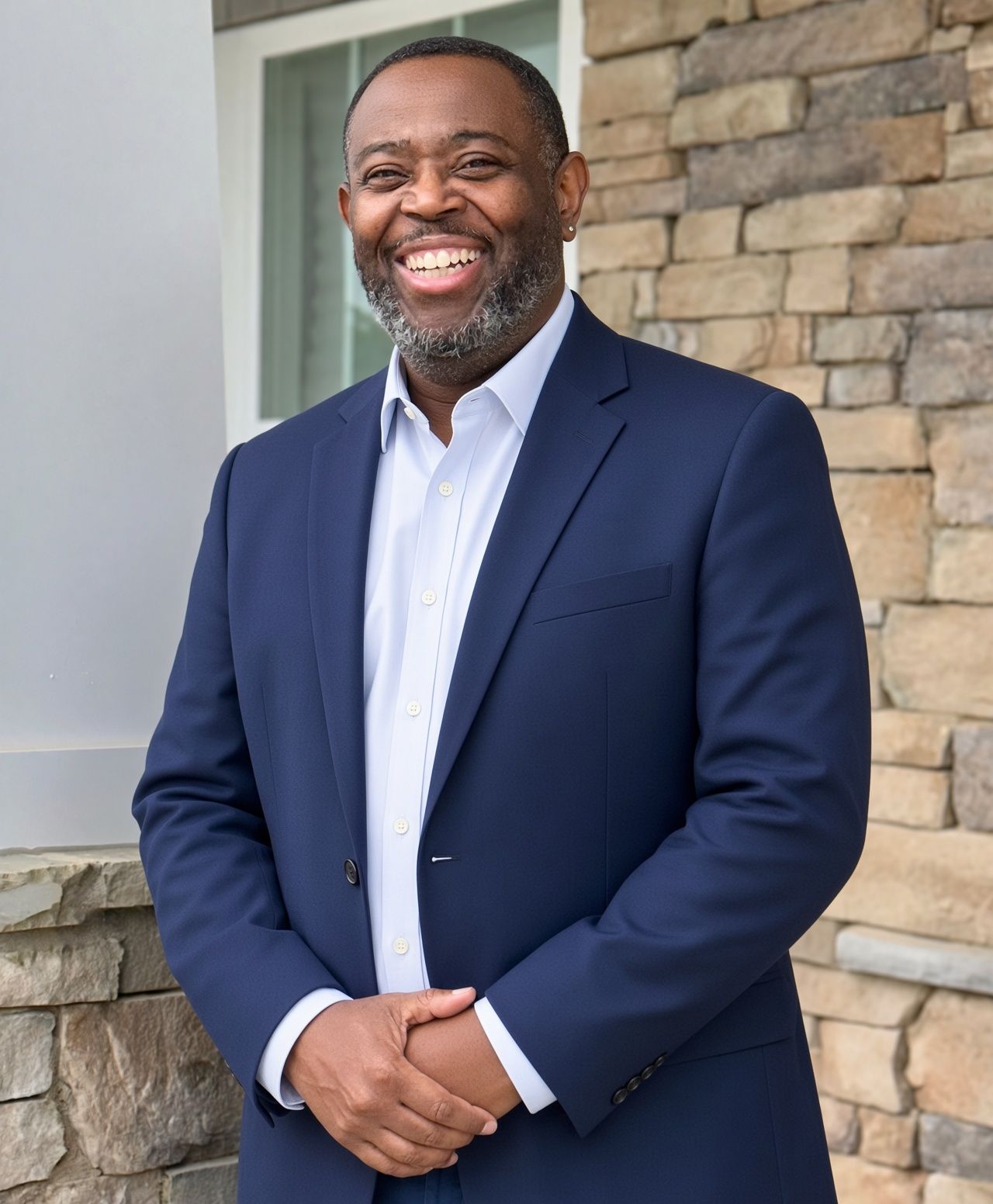 Man in a navy suit smiling beside a stone wall outdoors