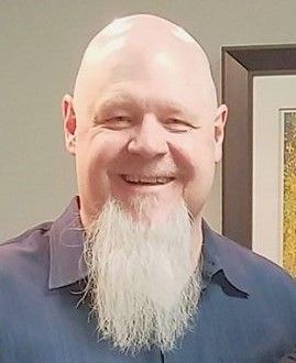 Smiling bald man with a long white goatee wearing a dark blue shirt indoors