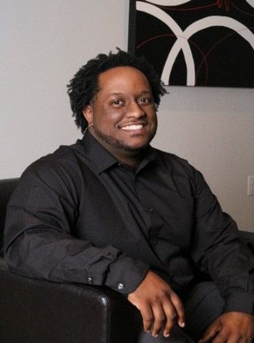 Smiling person in a black shirt seated in a dark armchair indoors with abstract wall art behind them