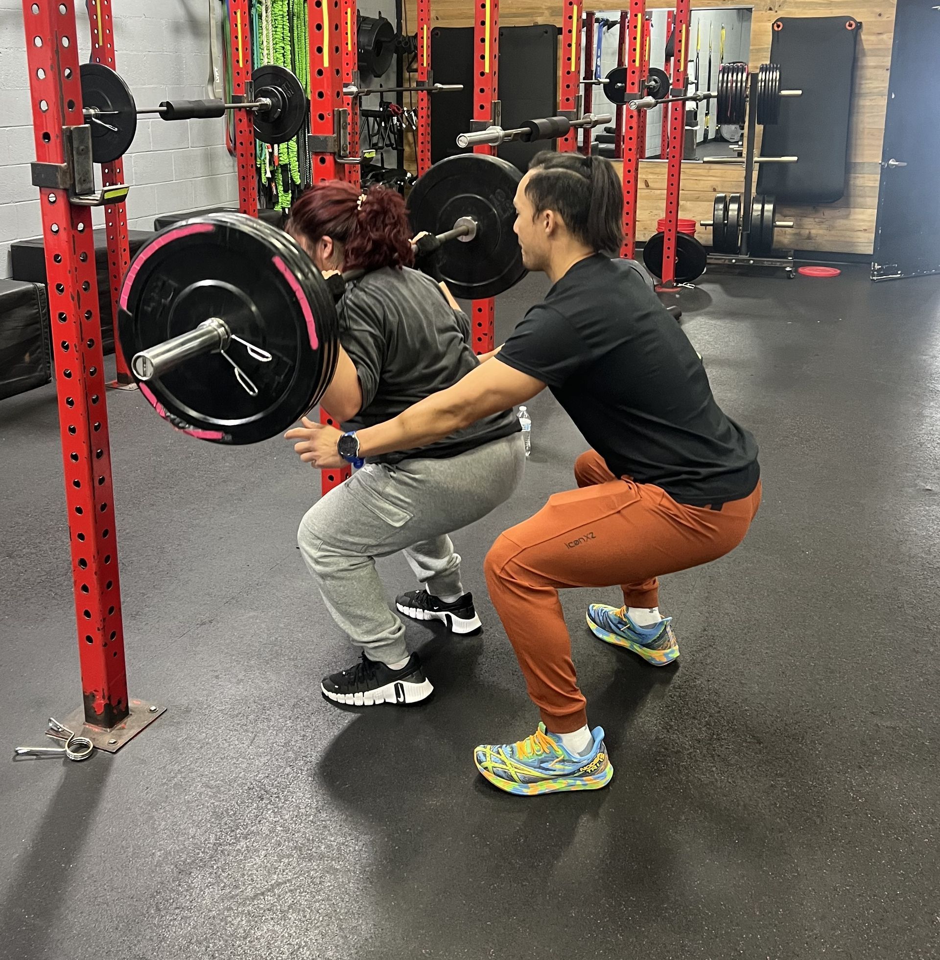 Man bench presses weight with a spotter at Team West Fitness, a gym in Westminster CA. Red and black weights and equipment.