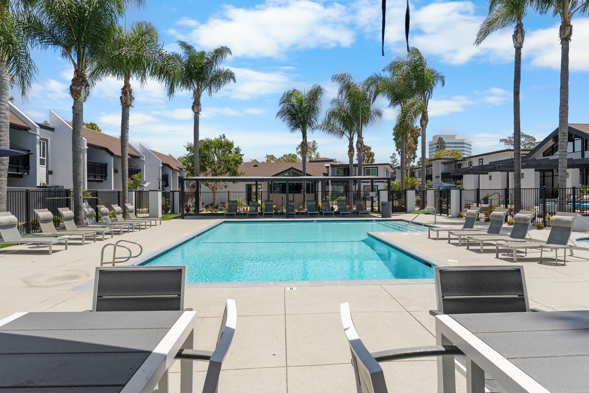 A large swimming pool is surrounded by palm trees and tables and chairs.